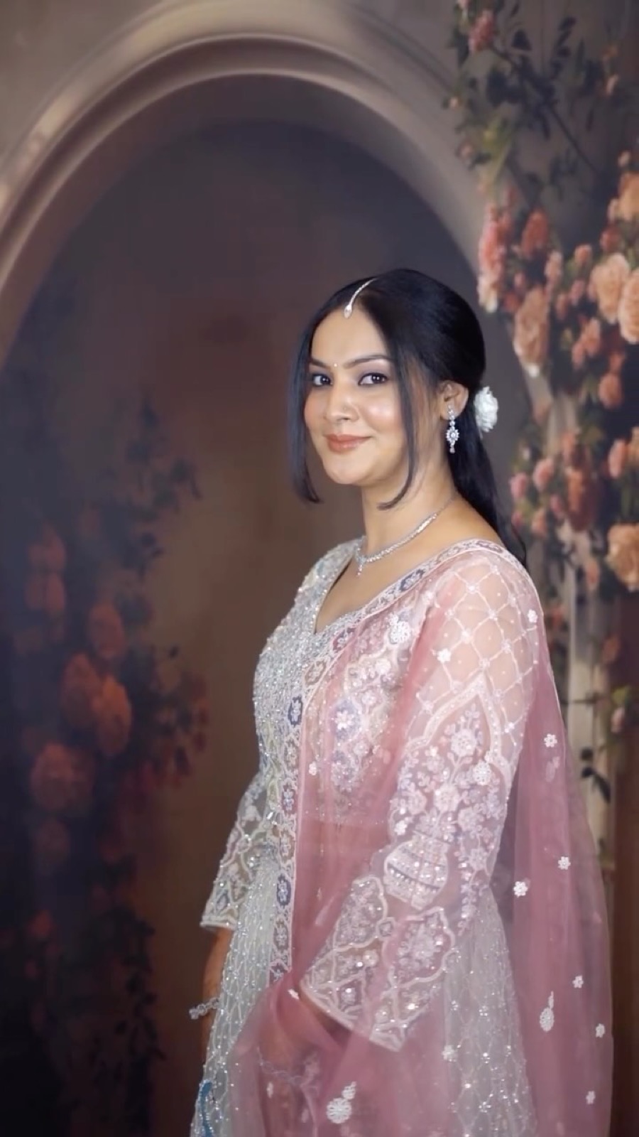 Bride in a pearl-embroidered suit with rose dupatta against a floral arch, makeup by Fashion World Palampur